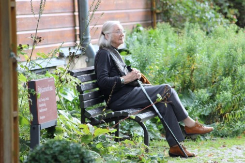 Ältere Frau mit Stock sitzt auf einer Bank im Grünen neben Haus